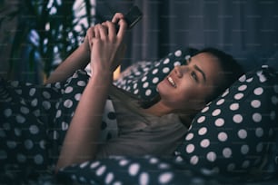 Bella ragazza sdraiata a letto e usando lo smartphone per chattare