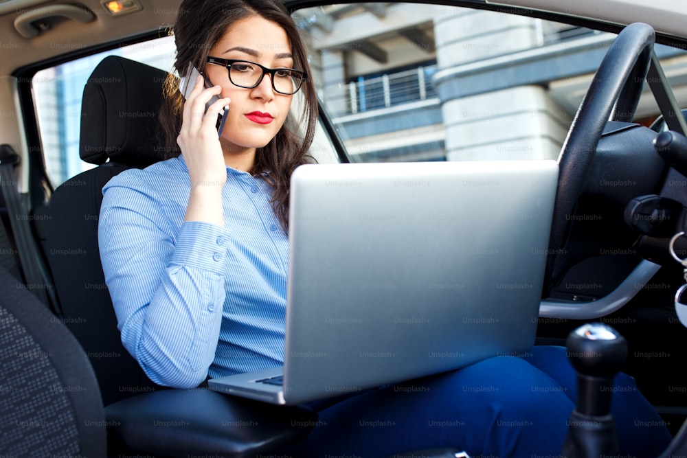 Beautiful young business woman sitting in the car with laptop and talking at phone.