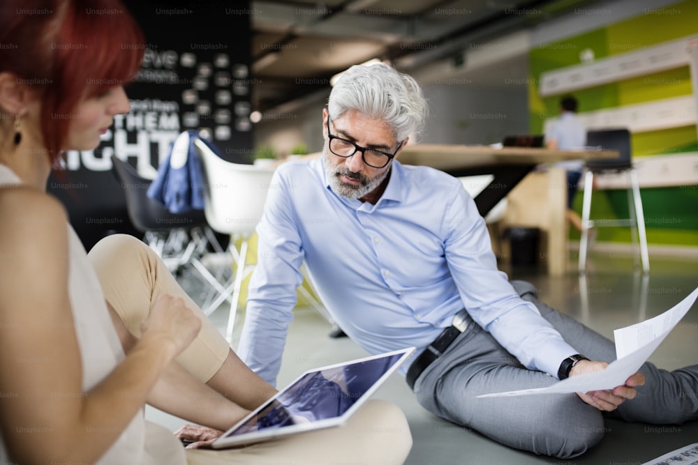 Duas pessoas de negócios com tablet no local de trabalho. Mulher e homem sentados no chão do escritório consultando um projeto juntos.