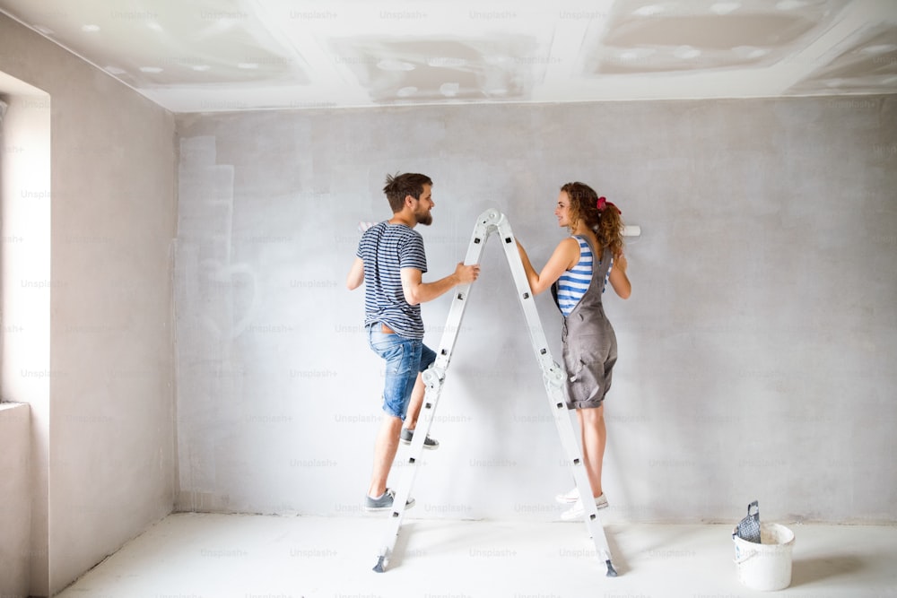 Beautiful young couple at home standing on ladder painting walls in their new house using paint rollers. Home makeover and renovation concept.