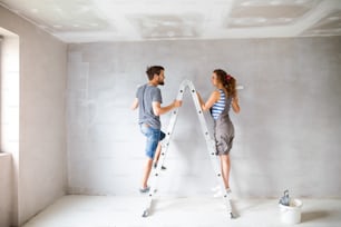 Beautiful young couple at home standing on ladder painting walls in their new house using paint rollers. Home makeover and renovation concept.