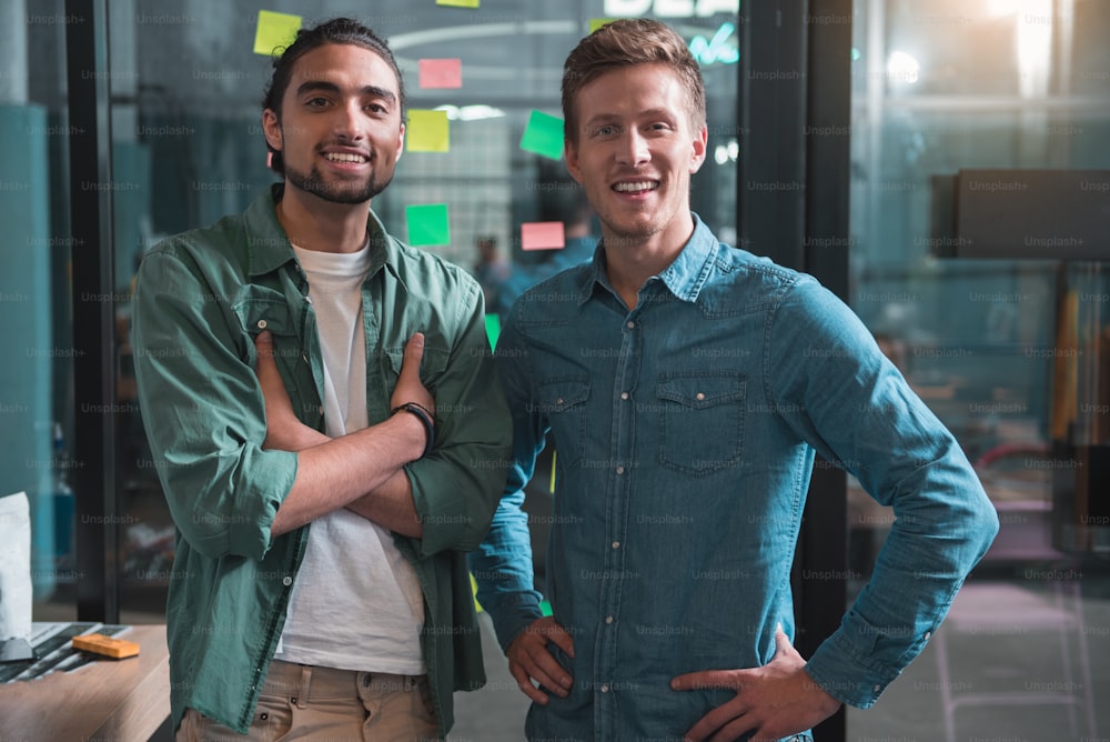 We are successful team. Portrait of confident young colleagues are standing against glass wall with stickers while looking at camera with joy. They are expressing gladness while laboring together