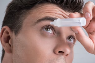 Pflege der Sehkraft. Nahaufnahme des Gesichts des jungen angenehmen Mannes verwendet medizinisches Produkt für seinen Augapfel. Isolierter Hintergrund