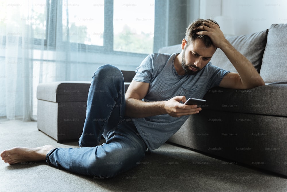 Comunicazione online. Uomo bello triste e triste seduto sul pavimento e con in mano il suo smartphone mentre controlla i messaggi