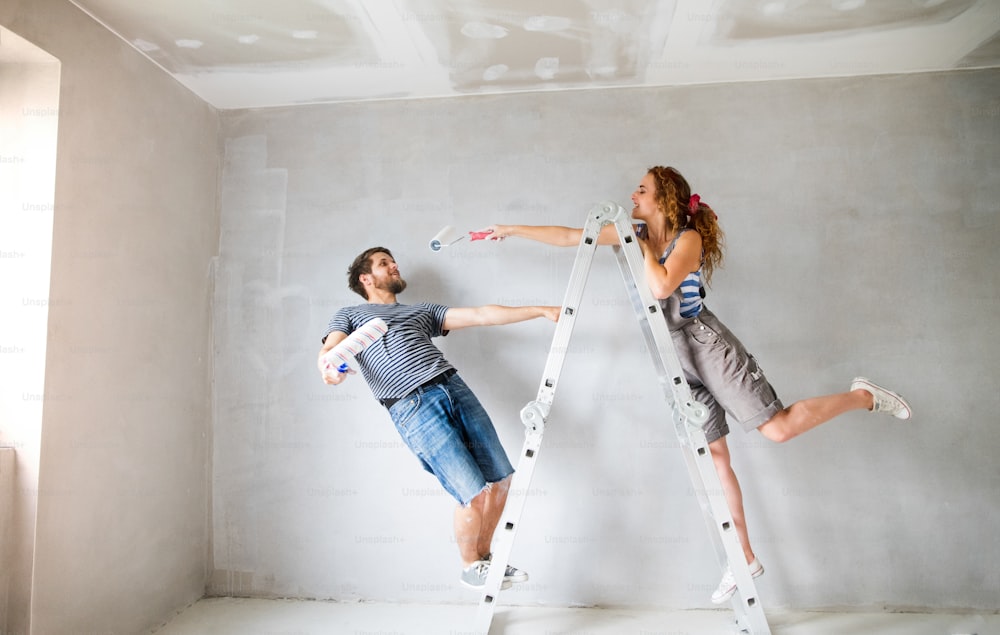 Beautiful young couple at home standing on ladder painting walls in their new house using paint rollers. Home makeover and renovation concept.