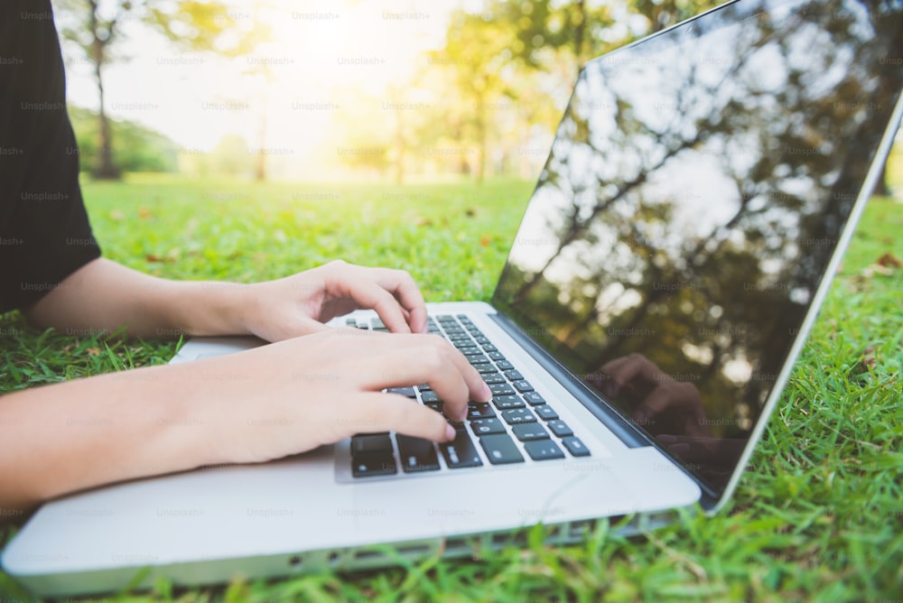 Junge Asiatin auf dem grünen Gras mit offenem Laptop. Mädchenhände auf der Tastatur. Fernstudienkonzept. Glückliche Hipster-junge asiatische Frau, die im Park am Laptop arbeitet. Student studiert im Freien.