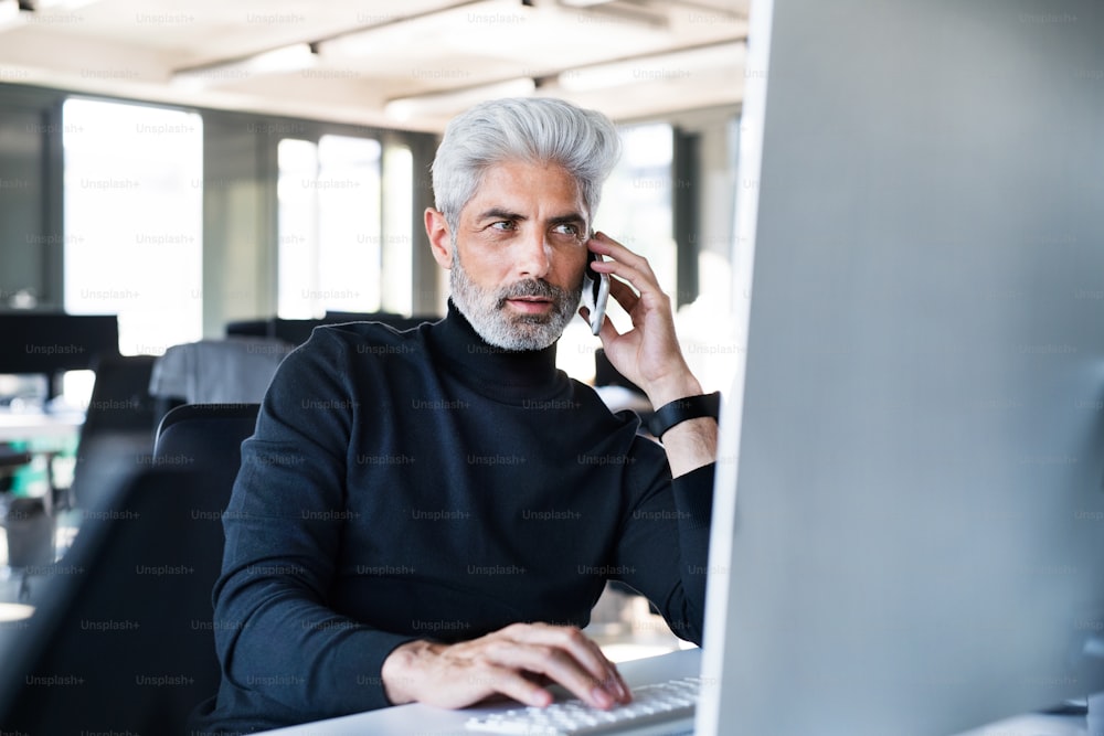 Hombre de negocios maduro con computadora portátil y teléfono inteligente sentado en el escritorio de la oficina, haciendo una llamada telefónica.