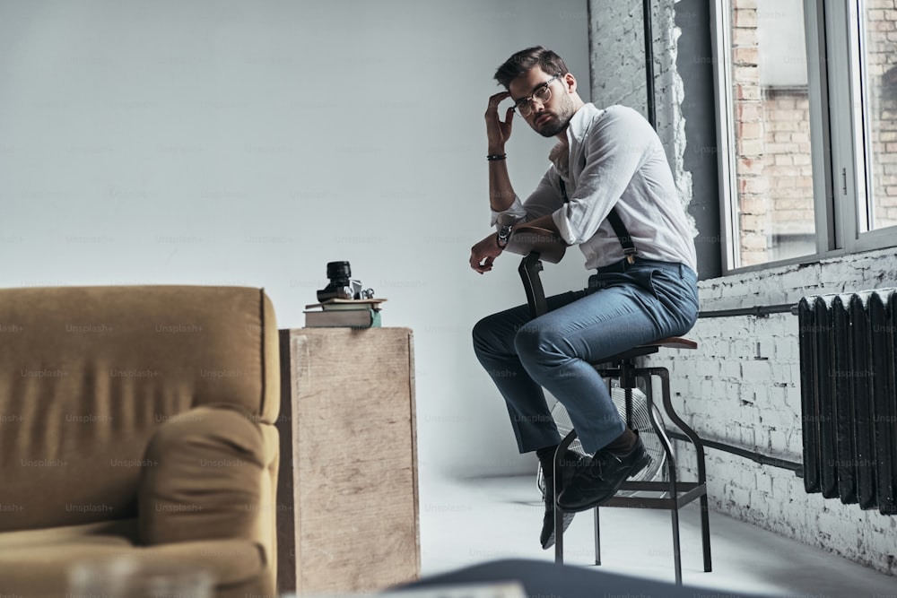 Handsome young man looking at camera and touching his head with hand while sitting on stool