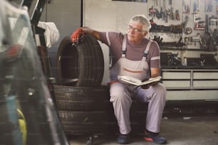 Senior man in workshop. Man sitting on tires and holding his notes.