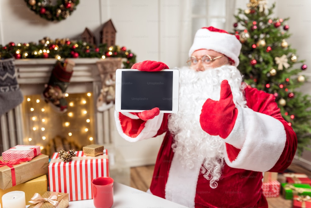 Best present ever. Cheerful old bearded man in costume is presenting tablet and giving thumb up. He is sitting at table near Christmas present boxes. Focus on gadget