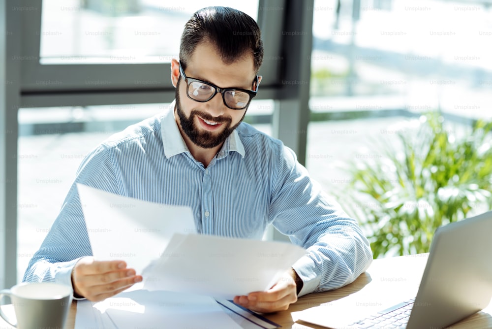 Careful inspection. Upbeat pleasant bearded man in glasses sitting in the office and looking through the documents in his hands