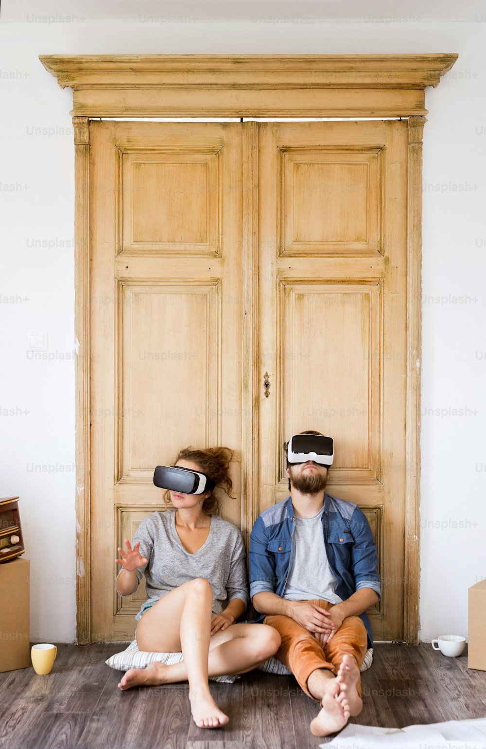Young married couple moving in new house, sitting on the floor, wearing virtual reality goggles.
