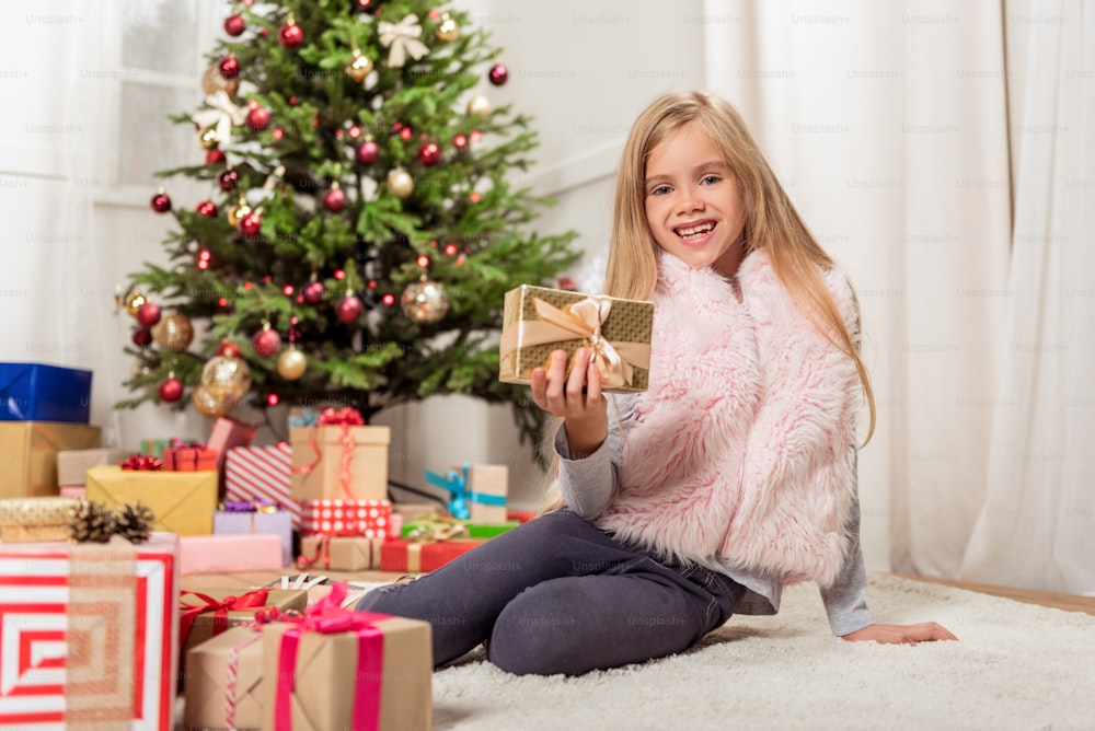 Ich mag Weihnachten. Porträt eines glücklichen Mädchens, das eine verpackte Geschenkbox vor der Kamera zeigt und lächelt. Sie sitzt auf dem Boden in der Nähe von vielen Kisten mit Tannenbaum im Hintergrund