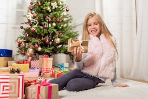 I like Christmas. Portrait of happy girl showing wrapped gift box to camera and smiling. She is sitting on floor near lots of boxes with fir-tree on background