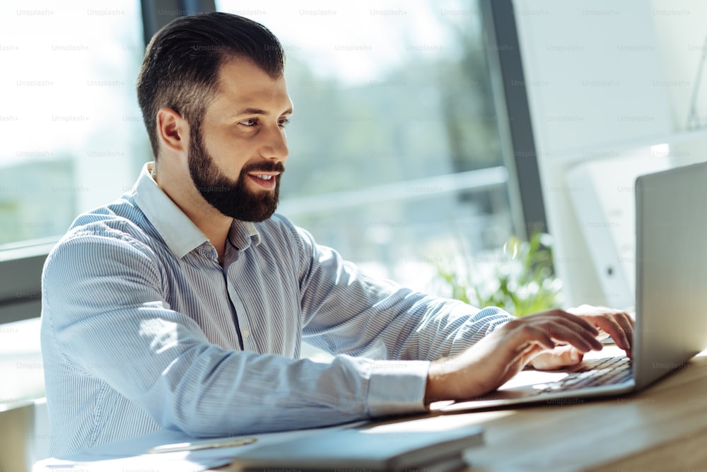 New ideas. Handsome bearded man sitting in the office and typing on the laptop, putting down his ideas, while smiling