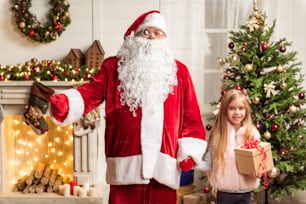 We love Christmas. Portrait of joyful fat Santa Claus and little child holding hands. They are standing near Christmas tree and fireplace at home. Girl is holding present and smiling
