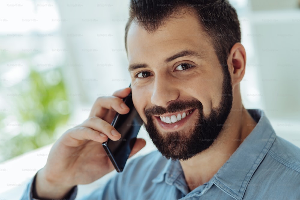 Charming smile. The close up of a cheerful handsome man talking on the phone and smiling at the camera broadly