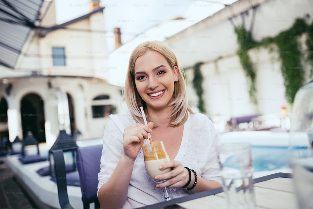 Young and beautiful blonde woman holding drink, smiling and looking at camera. Private house with swimming pool in background.