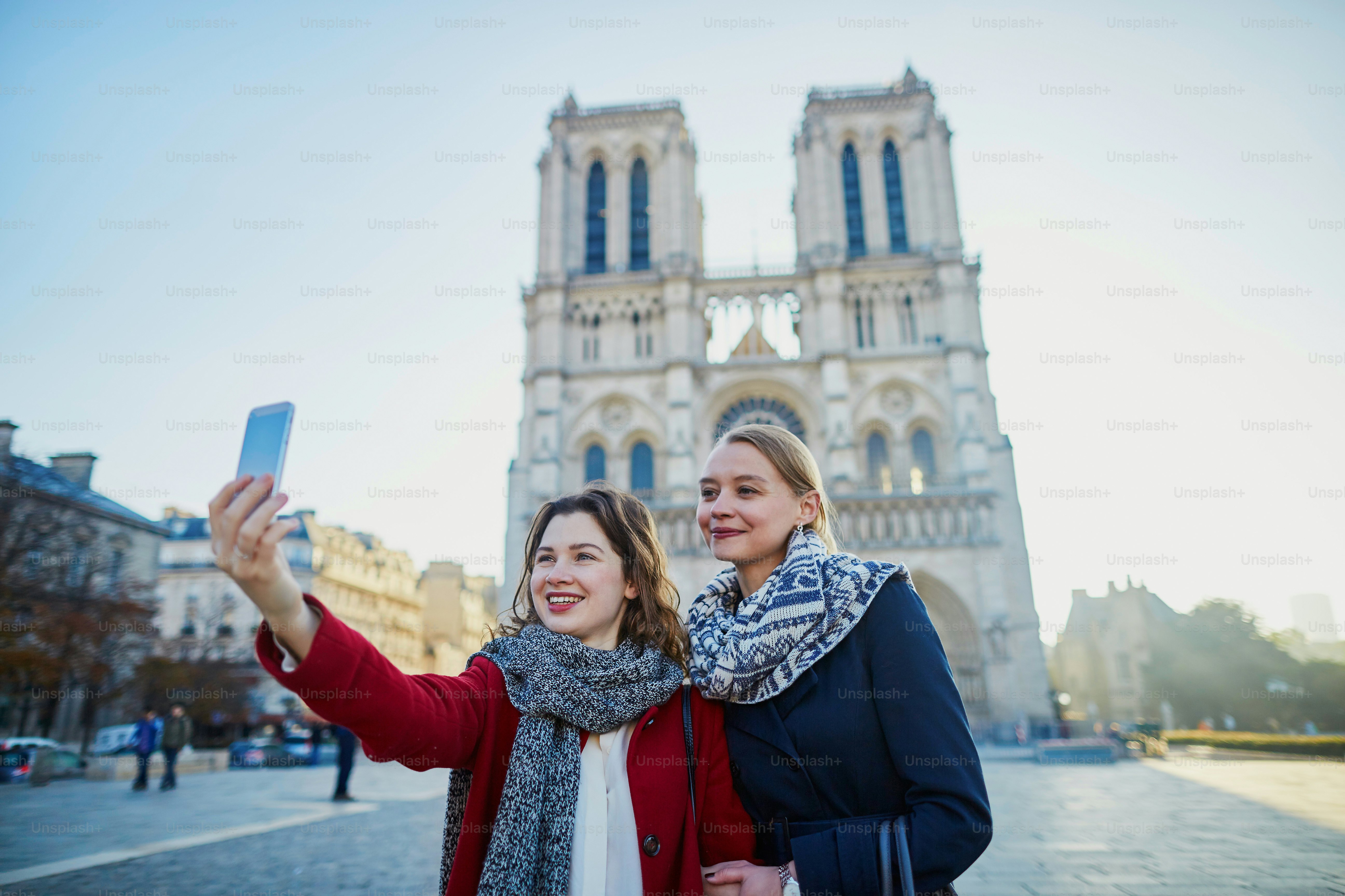 Two young girls walking together in Paris taking selfie with mobile phone near Notre-Dame cathedral. Tourism or friendship concept
