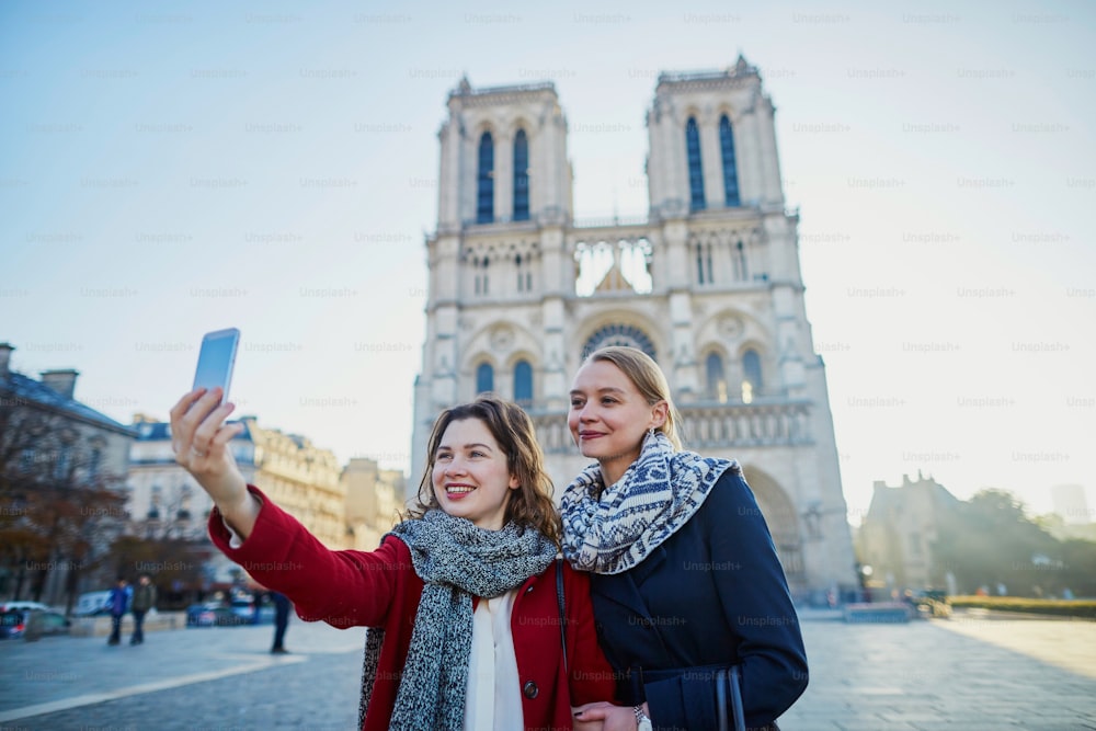 Two young girls walking together in Paris taking selfie with mobile phone near Notre-Dame cathedral. Tourism or friendship concept