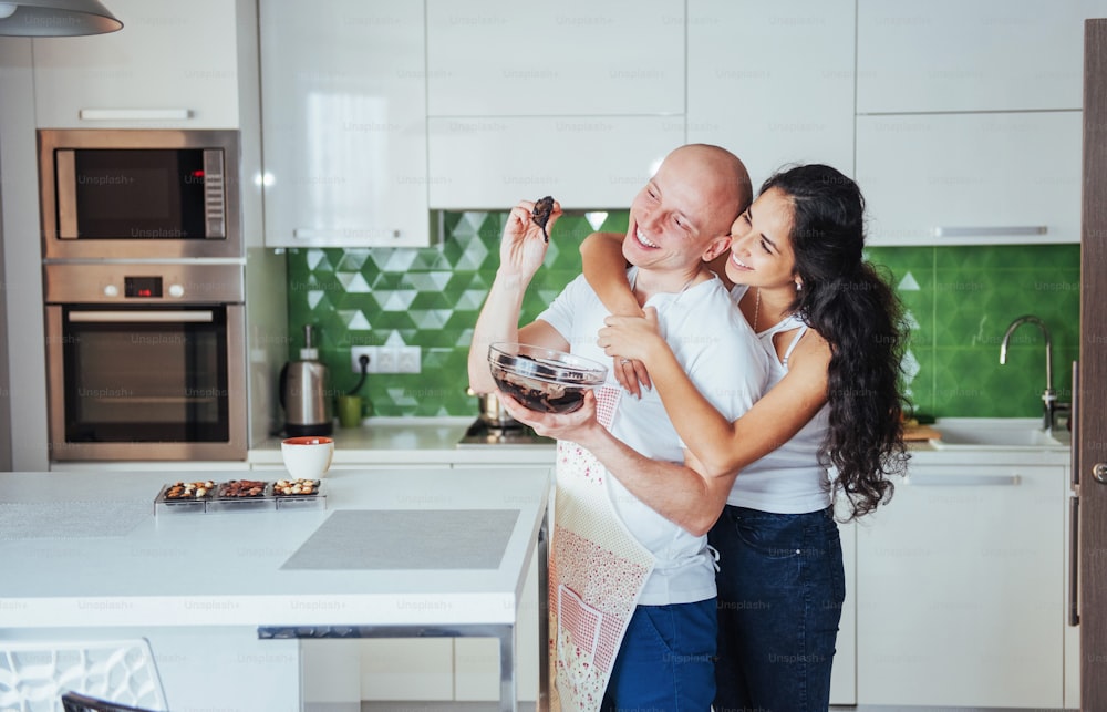 Beautiful young couple is talking, looking at camera and smiling while cooking in kitchen at home.