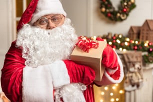 Feliz Navidad. Retrato del amable Padre Escarcha mostrando la caja de regalos y sonriendo. Lleva un traje rojo y blanco