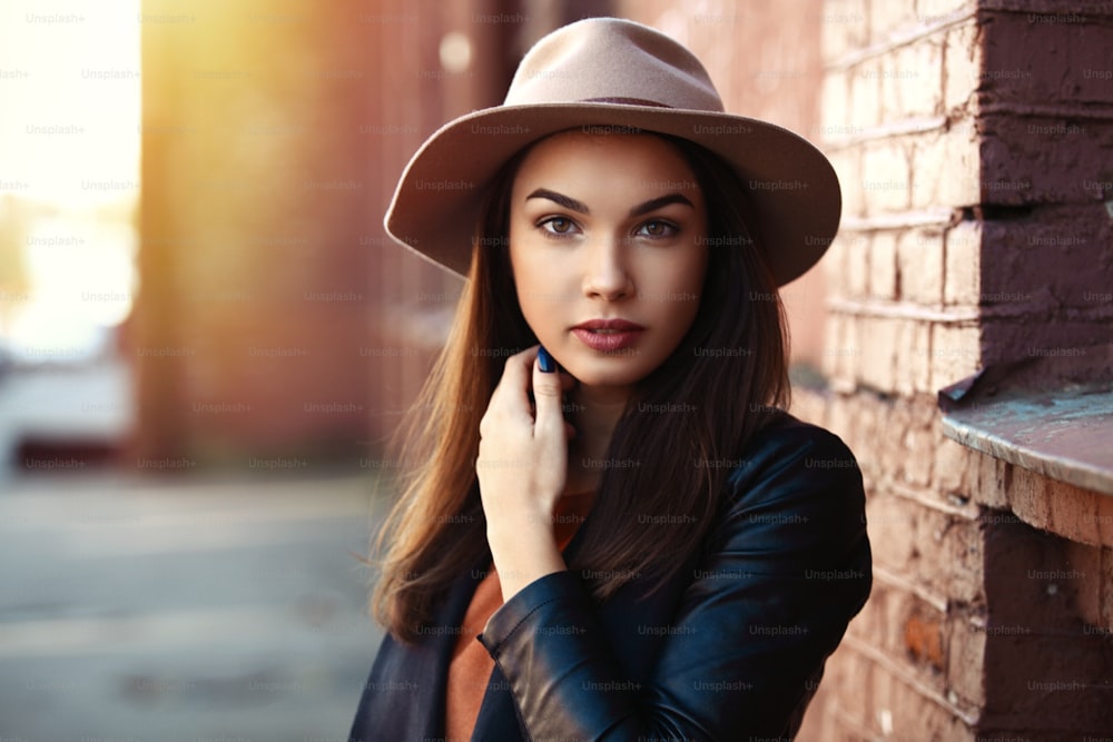 Portrait de femme de mode en gros plan de jeune jolie fille branchée posant à la ville, mode de rue d’automne, tenant un chapeau fedora rétro populaire jusqu’aux années 60. riant et souriant portrait.trendy