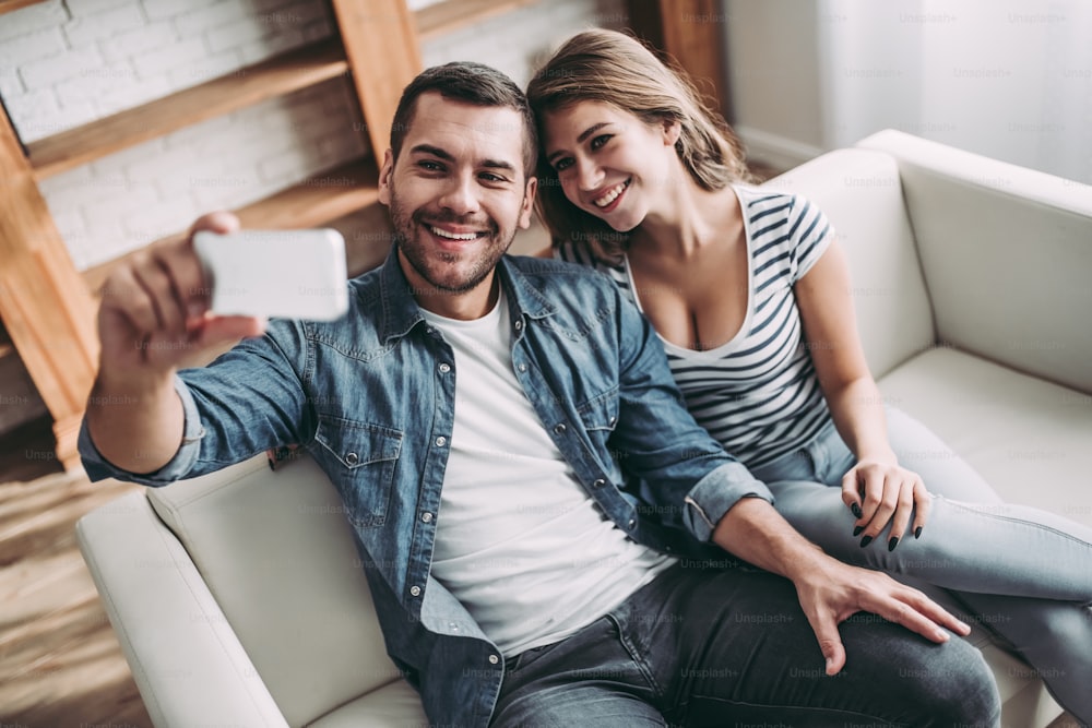 幸せなカップルは自宅のソファに座っています。抱きしめたり、笑顔で、スマートフォンで自撮りをしたり。