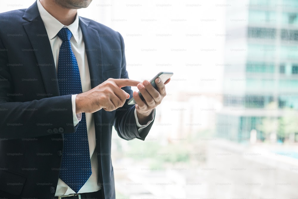Homme d’affaires utilisant une application de téléphone portable envoyant des SMS à l’extérieur du bureau dans une ville urbaine avec des gratte-ciel en arrière-plan. Jeune homme asiatique tenant un smartphone pour le travail professionnel.