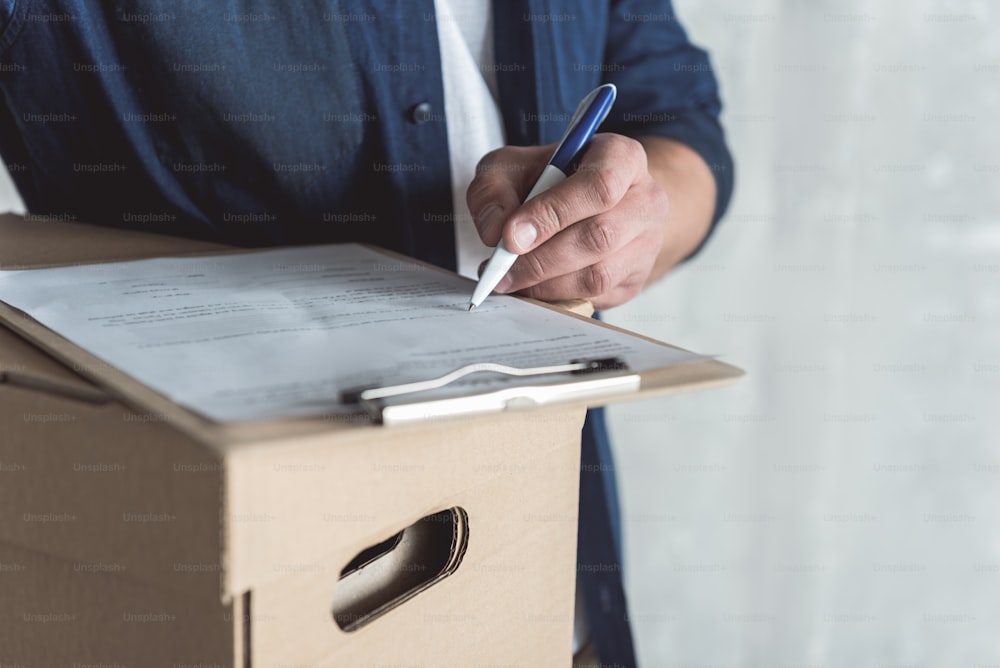 Place for sign. Close-up of hand with pen of professional courier who is holding cardboard box and folder with document. He is putting his signature while delivering personal order