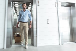I like traveling. Full length portrait of delightful stylish businessman with stubble is exiting from modern lift while carrying his luggage. He is looking aside with smile. Copy space in the right side