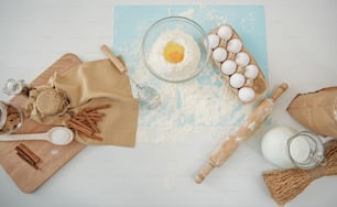 Top view of broken egg on flour in bowl, rolling pin and milk, wooden board with cookies and cinnamon on the table