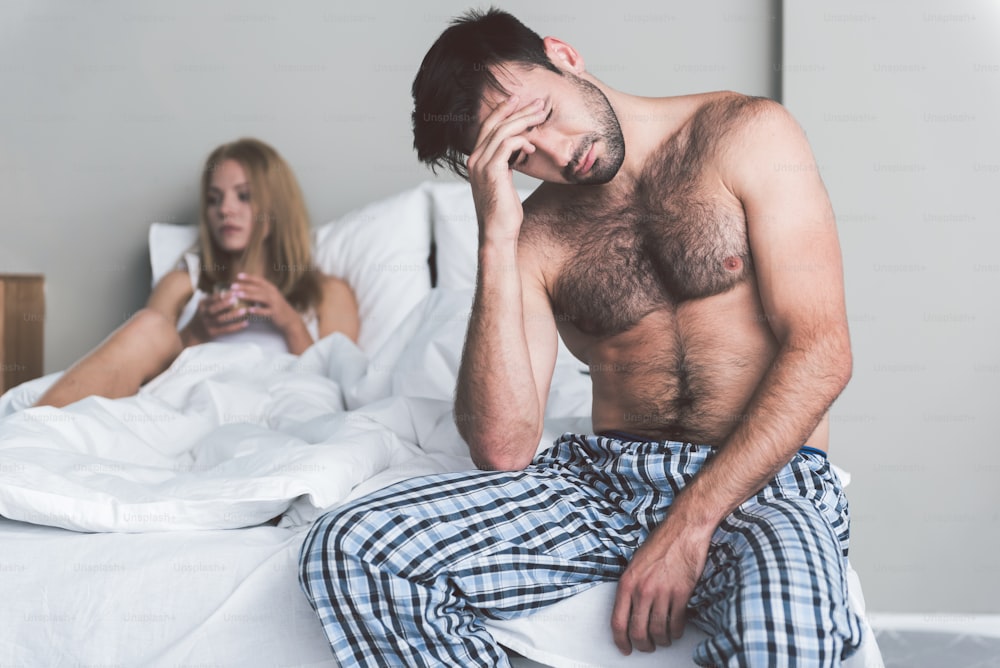 Family conflict. Tired young man is touching his head and closed eyes with desperation. Sad woman is lying in bed on background