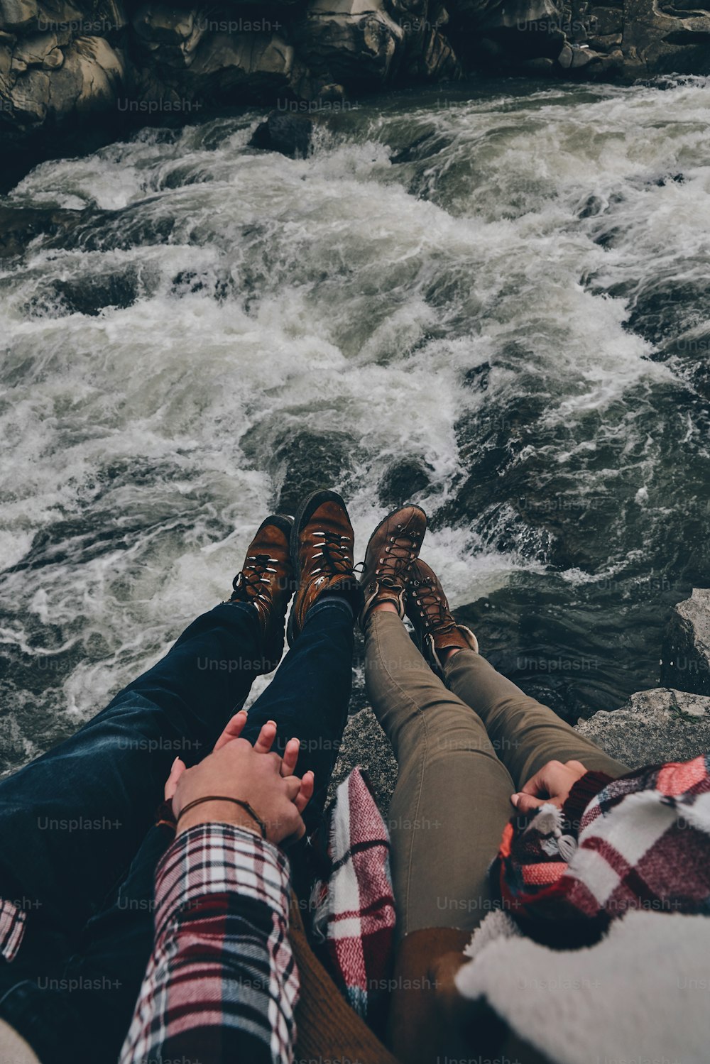 Close up top view of young couple holding hands while sitting on the rocks with the river below