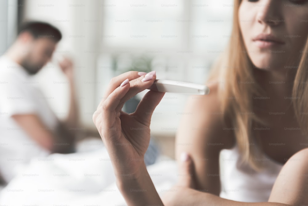 Pensive young woman is checking result on her pregnancy test. Focus on stick in her hand. Nervous man is sitting on background