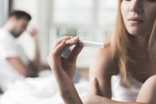 Pensive young woman is checking result on her pregnancy test. Focus on stick in her hand. Nervous man is sitting on background