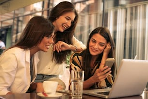 Three young woman sitting in cafe using smart phone and having funny conversation. One of them watching on clock.