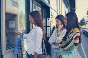 Young woman with friends take money from cash machine. She using credit card. Her friends watching and waiting .