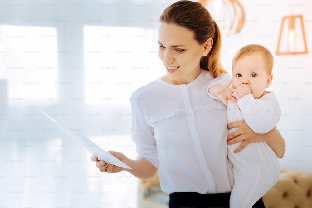 Documents curieux. Jeune femme positive et attentive lisant les documents tout en tenant un bébé dans ses bras