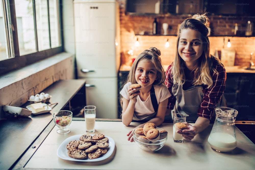 Attractive young woman with her little cute daughter are cooking on kitchen. Drinking milk, eating cakes and cookies.