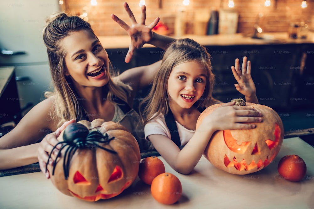 Feliz Dia das Bruxas! Jovem atraente com sua pequena filha fofa estão se preparando para o Halloween na cozinha. Mamãe com filha estão se divertindo com abóboras.