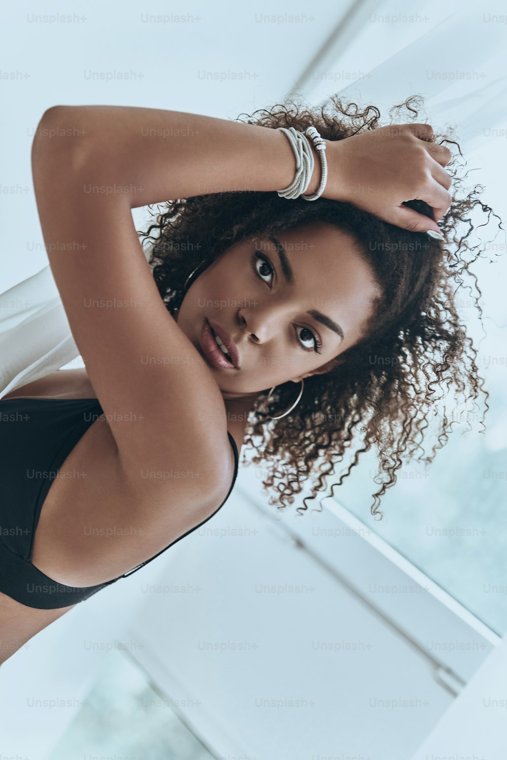 Attractive young African woman in black lingerie keeping hand in hair and looking at camera while standing near the window at home