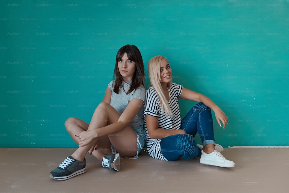 Two young beautiful girls enjoy each others company siting together on a turquoise background, crossed legs, looking to camera. Friendship, beauty, best friends.