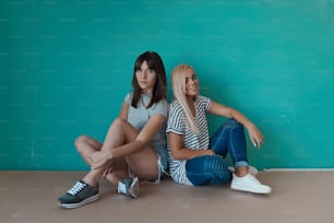 두 명의 젊은 아름다운 소녀는 청록색 배경에 함께 앉아 다리를 꼬고 카메라를 바라보며 서로의 회사를 즐깁니다. 우정, 아름다움, 가장 친한 친구.