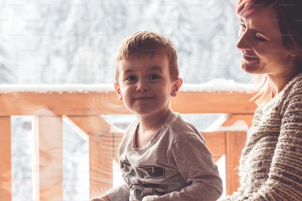 Mother and her son sitting in a comfortable chair next to the window with look at snow covered mountain.