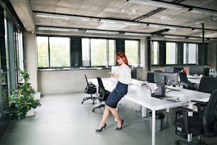 Schöne junge Geschäftsfrau mit Smartphone in ihrem Büro, die auf dem Schreibtisch sitzt und eine Pause macht.