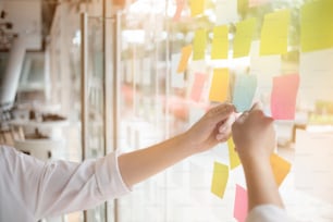 Les gens d’affaires créatifs lisant des notes autocollantes sur un mur de verre avec un collègue de travail utilisent des notes post-it pour partager des idées en discutant et en travaillant en équipe, en faisant du brainstorming. Plan rapproché.