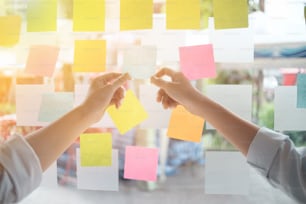 Les gens d’affaires créatifs lisant des notes autocollantes sur un mur de verre avec un collègue de travail utilisent des notes post-it pour partager des idées en discutant et en travaillant en équipe, en faisant du brainstorming. Plan rapproché.
