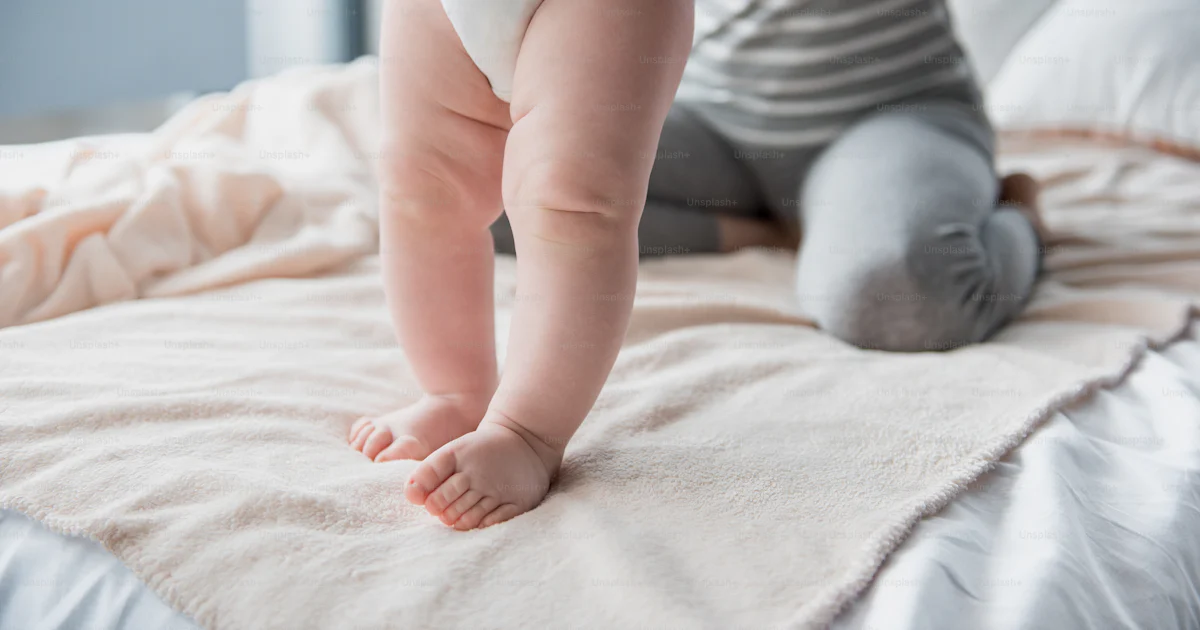 Close up of infant legs standing on bed, female sitting on background ...
