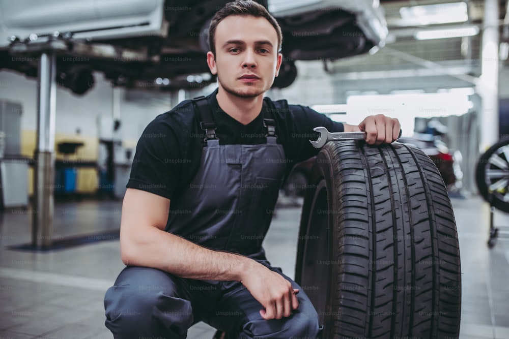 Handsome mechanic in uniform is working in auto service. Car repair and maintenance. Holding car wheel/tire.